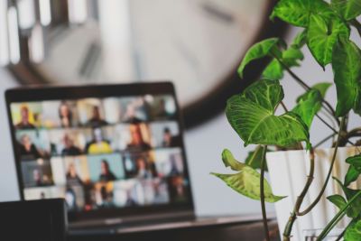 A laptop showing a multi‑person video call on screen, positioned behind a green plant, representing a virtual meeting or online webinar environment.